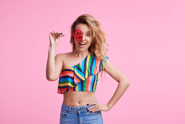 Sweet and candy. The girl colorful dress on pink background in the studio. Woman holding a red heart-shaped lollipop. Valentine's Day. Girl holding a lollipop in open mouth. Wow and shocked concept.  - Foto, imagen