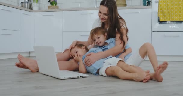 Moeder met twee jonge kinderen, zittend op de vloer in een moderne appartement in een lichte interieur met een laptop. Bekijk de laptop scherm en bekijk familiefoto's. Online aankopen doen voor kinderen. - Video