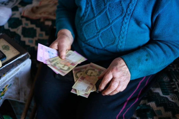 Yaşlı, gri saçlı kadın Ukrayna 'nın kağıt parasını elinde tutuyor. Kadın üzgün. Köyde fakir bir hayat. Yaşlılık iyi değil. Düşük ışık fotoğrafı. - Fotoğraf, Görsel