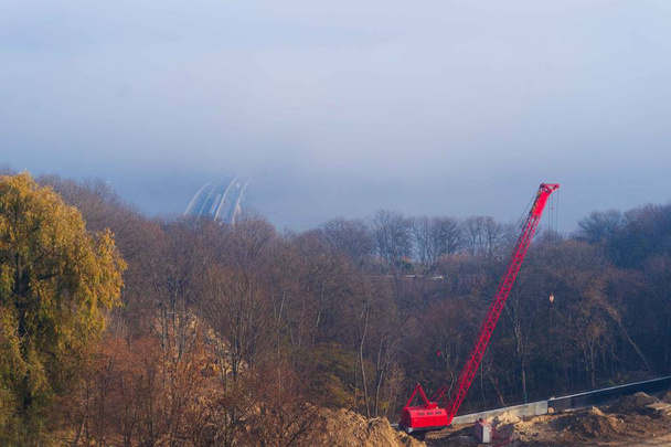 赤いギベットと建設自動車クレーンと建設プロセス。背景に地下鉄の列車と秋の霧と川の橋。鶴の周りの森. - 写真・画像
