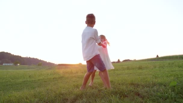 SLOW MOTION LENS FLARE: Cheerful kids holding hands and dancing at golden sunset. Playful brother and sister playing ring around the Rosie in meadow field. Lively boy and girl having fun in summer - Footage, Video