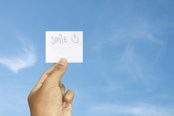 Χέρι κρατώντας το χαμόγελο λέξη σε χάρτινη κάρτα με μπλε ουρανό φόντο. - Φωτογραφία, εικόνα