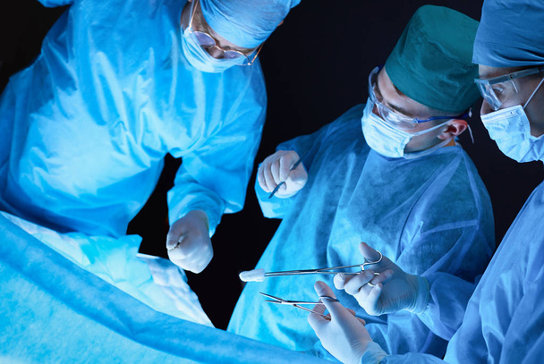 Group of surgeons at work in operating theater toned in blue. Medical team performing operation - 写真・画像