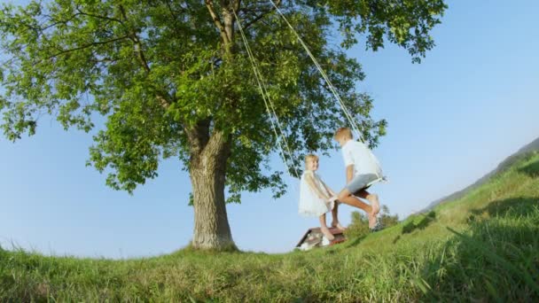 LOW ANGLE VW Smiling мальчик и девочка наслаждаются теплым солнечным днем, качаясь под большим деревом. Брат и сестра веселятся на открытых качелях. Босоногие счастливые дети, качающиеся на качелях, свисающих с дерева
 - Кадры, видео