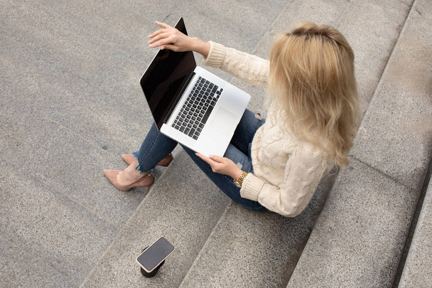 Young pretty woman sitting with laptop having coffee break on the stairs. Caucasian girl working with notebook in the outdoors. - Foto, Imagen