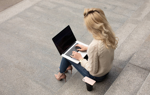 Young pretty woman sitting with laptop having coffee break on the stairs. Caucasian girl working with notebook in the outdoors. - Foto, Imagem
