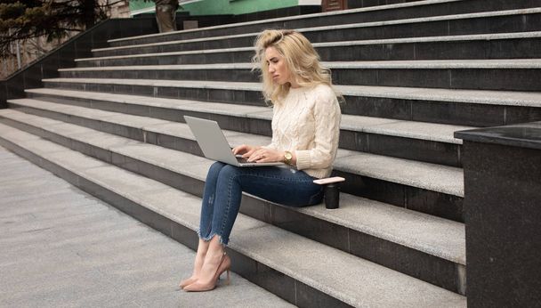 Young pretty woman sitting with laptop having coffee break on the stairs. Caucasian girl working with notebook in the outdoors. - Φωτογραφία, εικόνα