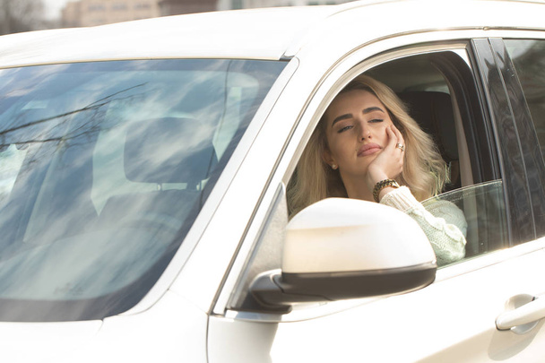 Beautiful woman is sitting and driving a car. She is holding a mobile phone and messaging. The lady is drinking coffee and looking forward happily. She is smiling - Zdjęcie, obraz