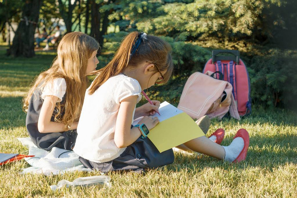 Dos amigas colegialas aprendiendo sentadas en un prado en el parque. Niños con mochilas, libros, cuadernos. Vista desde atrás
 - Foto, Imagen