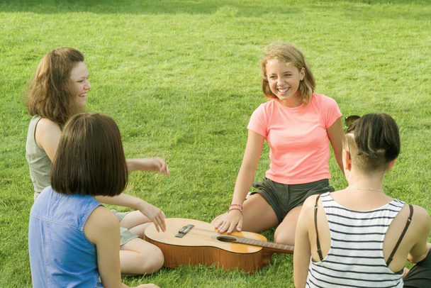 Group of happy teenage girls having fun outdoors with guitar. Come up with new music, sit on green lawn in the yard - 写真・画像
