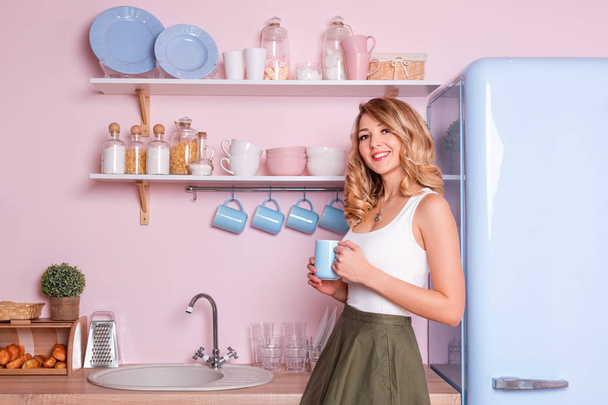 Young happy woman drinking coffee or tea at home in the kitchen. Blonde beautiful girl having her breakfast before going to office. Coffee break. Pink and blue pastel modern kitchen interior - Φωτογραφία, εικόνα