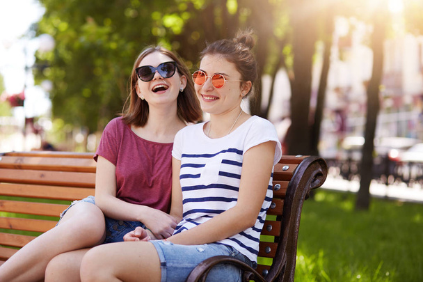 Porträt zweier attraktiver junger Freunde, die sich nach einem langen Spaziergang auf einer Holzbank im örtlichen Park ausruhen. lachende Mädchen haben jede Menge Energie, um ihren Spaß fortzusetzen. Konzept der Freundschaftsbeziehungen. - Foto, Bild