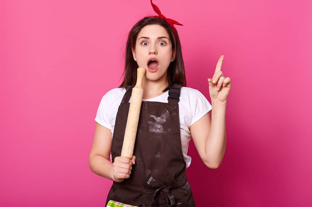Busy beautiful cook opens her mouth widely, raise her finger up, holds rolling pin, wears cooking uniform and bright red headband. Emotional slender woman has new cool idea of delicious recipe. - Fotografie, Obrázek