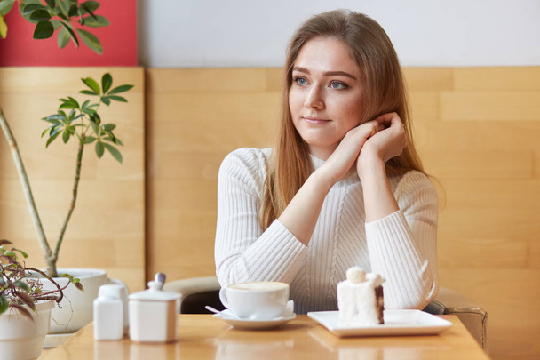 Beautiful model sits in local cafe, looks in window, eats delicious cake and drinks coffee, puts her head on hands, feels satisfied. Tender young girl having rest in cosy and pleasant atmosphere. - Fotoğraf, Görsel