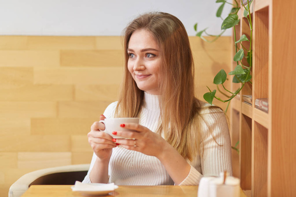 Sincere cute young lady sits in local cafeteria near wooden shelf with green plant behind her, holds small white cup of coffee in both hands, looks in other way, warms up in cosy public place. - Photo, image