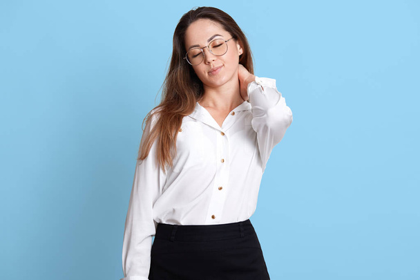 Portrait of pleased smiling young businesswoman in glasses, isolated over blue background, massages her stiff neck with one hand after stressful day, feels relaxed. Copy space advertisement. - Foto, Imagem