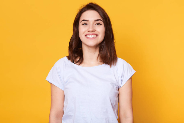 Portrait of emotive good looking caucasian woman laughs while looking directly at camera and standing against yellow background. Charming brunette female with toothy smile looks happy, has great news. - Photo, Image