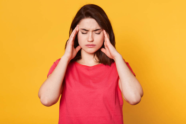 Stressed beautiful woman wears casual t shirt, touching temples and has terrible headache, attractive young lady looks tired. Stress or headache concept. Model posing isolated over yellow background. - Foto, imagen