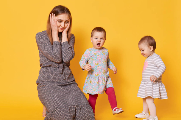A portrait of stressed tired mother who covers her ears with two hands from kid`s loud shouting, suffers from awful headache, has playful disobedient children. Upbringing children concept. - Foto, imagen