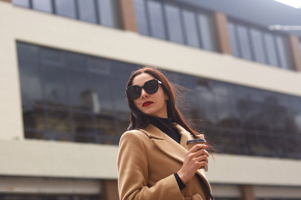 Portrait of calm elegant woman turning and looking over shoulder, dressed black sweater, beige coat and stylish sunglasses, holds takeaway coffee in hand, poses isolated over blurred street background - 写真・画像
