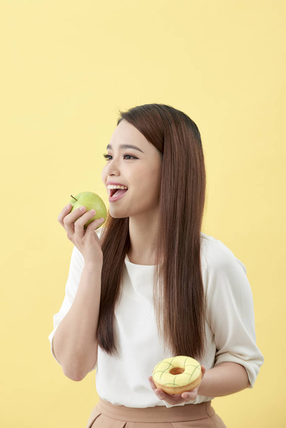 Elija una opción correcta para una buena salud. Las mujeres está eligiendo la opción entre donut y manzana verde aislado sobre amarillo
. - Foto, imagen