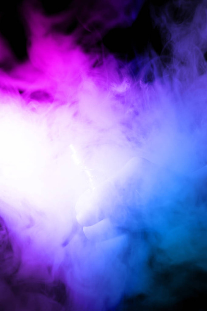Männliche Hand hält Vape in dichtem Nebel oder Rauch. Vapowolken um die Hand mit Vape auf schwarzem Hintergrund. Nebel ist 2 Farben blau und lila. Brühe isoliert bunten Rauch mit Sprühglycerin. - Foto, Bild