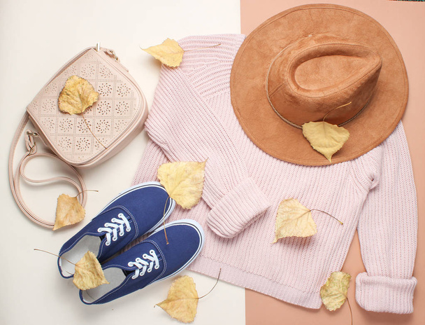 Women's clothing and accessories on a beige background. Fashionable sweater, sneakers, felt hat, leather bag among the fallen yellowed leaves. Autumn wardrobe. top view - Photo, Image