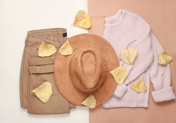 Γυναικεία ρούχα και αξεσουάρ σε μπεζ φόντο. Μοδάτο πουλόβερ, παντελόνια, καπέλο στα πεσμένα φύλλα. Φθινοπωρινή γκαρνταρόμπα. επάνω όψη. Επίπεδη ωοτοκίδα - Φωτογραφία, εικόνα