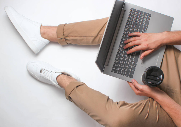 Töredéke a férfi lábak bézs nadrág és fehér cipők ül fehér. Ember használ modern laptop. Online munkás, freelancing, otthoni munka, dolgozó ember. felülnézet fehér háttérképet - Fotó, kép