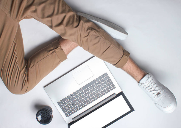 Fragment de jambes masculines en pantalon beige et baskets blanches assises sur blanc. Homme utilisant un ordinateur portable moderne. Travailleur en ligne, pigiste, travail à la maison, homme travaillant. vue de dessus sur fond blanc
 - Photo, image
