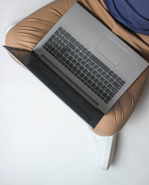 Fragment de jambes masculines en pantalon beige et baskets blanches assises sur blanc. Homme utilisant un ordinateur portable moderne. Travailleur en ligne, pigiste, travail à la maison, homme travaillant. vue de dessus sur fond blanc
 - Photo, image