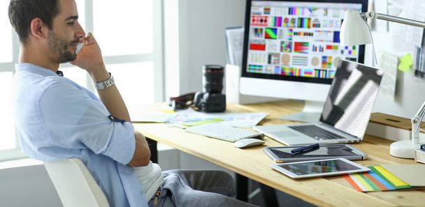 Πορτρέτο του νεαρού σχεδιαστή που κάθεται στο γραφιστικό στούντιο μπροστά από το laptop και τον υπολογιστή ενώ εργάζεται στο διαδίκτυο. - Φωτογραφία, εικόνα