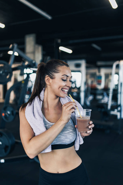 Young attractive woman after successful workout in modern fitness gym holding glass of protein shake and drinks with drinking straw. - Fotó, kép