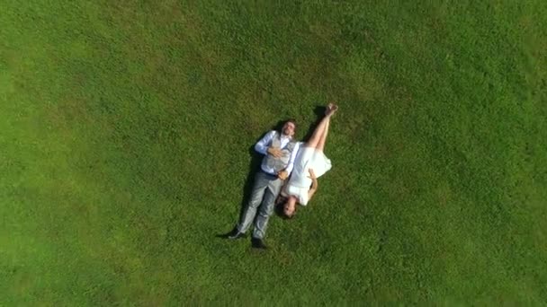 AERIAL TOP DOWN: Счастливая молодая пара лежит в траве в летний день. Мужчина и жена молодожены отдыхают в солнечном парке после свадьбы. Молодой человек и беременная женщина лгут и держатся за руки, ожидая ребенка
 - Кадры, видео