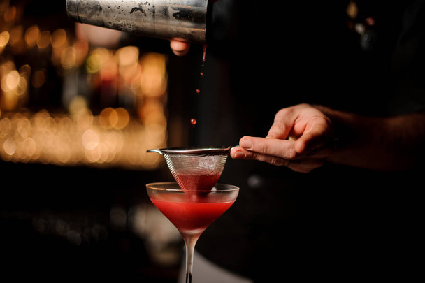 Professional bartender pouring a red cocktail through the sieve to the glass - Фото, изображение