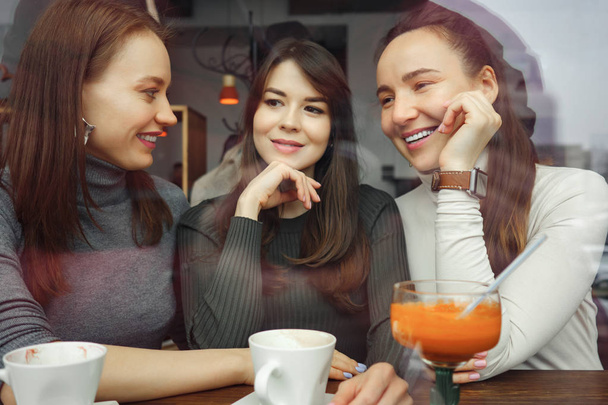 Las mujeres amigas en un café se están divirtiendo charlando y bebiendo sus bebidas. Vista desde detrás del vidrio
. - Foto, Imagen