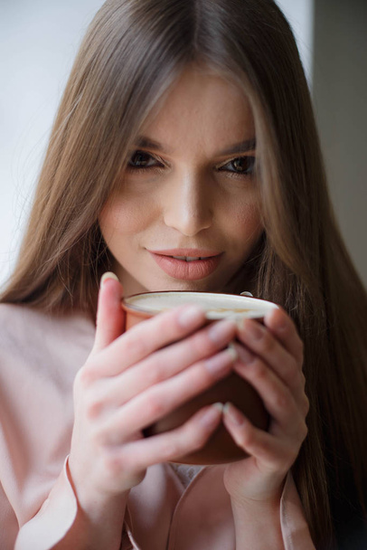 Beautiful girl is drinking coffee and smiling while sitting at the cafe - Zdjęcie, obraz