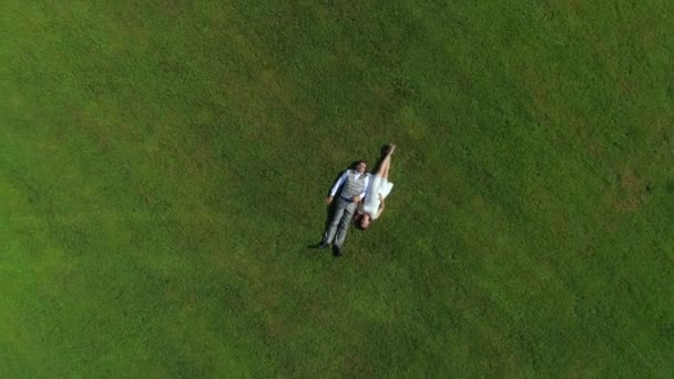 AERIAL TOP DOWN: Счастливая молодая пара лежит в траве в летний день. Мужчина и жена молодожены отдыхают в солнечном парке после свадьбы. Молодой человек и беременная женщина лгут и держатся за руки, ожидая ребенка
 - Кадры, видео