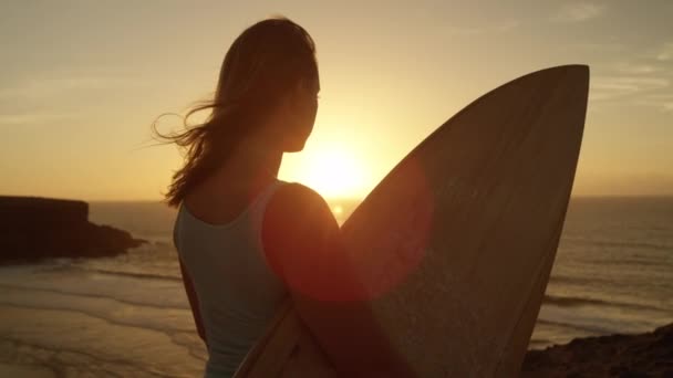 Повільний рух крупним планом: Фер дівчина стоїть на скелястому океані скелі і спостерігаючи розбиваючи хвилі на Золотий захід сонця. Жінка тримає дошку для серфінгу, дивлячись хвиль на світанку в вітряній Фуертевентура, Канарські острови - Кадри, відео