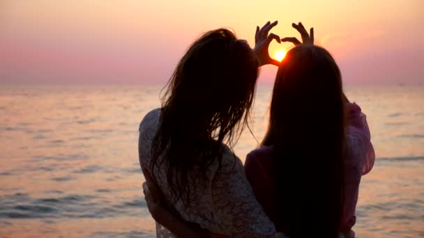 Lgbt のコンセプト。幸せなレズビアンのカップルは日没の美しいトロピカルビーチで休んでいます。女の子は抱きしめて、自分の手で心を作り、夕日に逆らって - 映像、動画