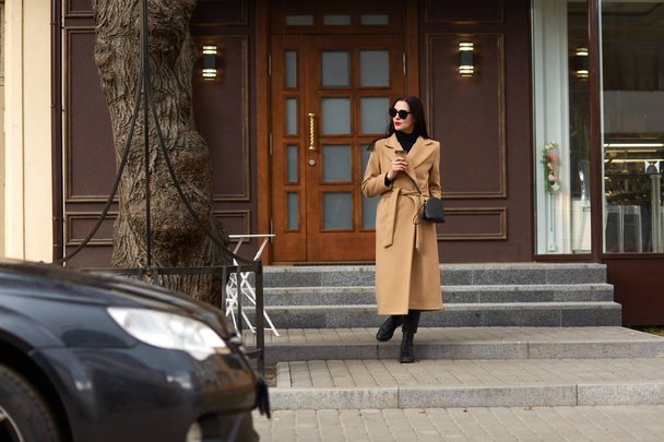 Full length photo of beautiful brunette female standing near city building, drinking hot takeaway coffee, going to her car, looks aside, wearing beige coat, boots, sunglasses, spends free time. - Fotó, kép