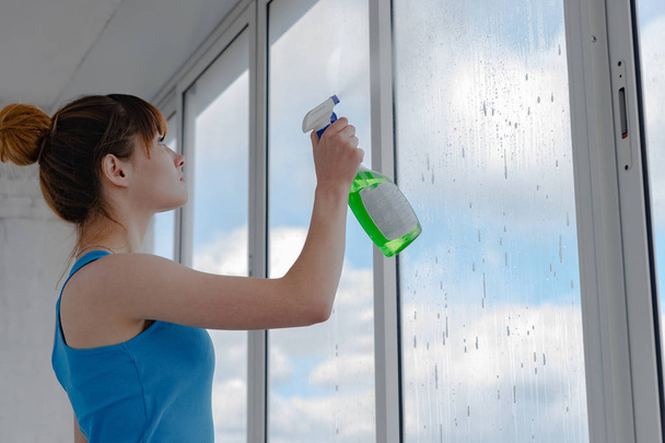 La ragazza spruzza il liquido per lavare finestre su vetro sporco. Una donna con una t-shirt blu lava una finestra
. - Foto, immagini