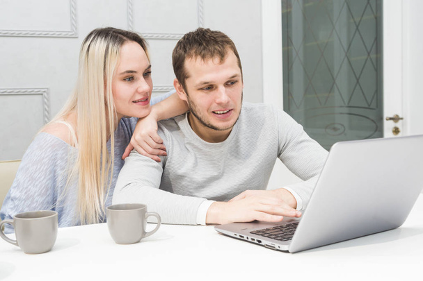 Young couple sitting at the morning in they kitchen and using laptop. Online shopping concept. Modern kitchen in background. - Foto, imagen