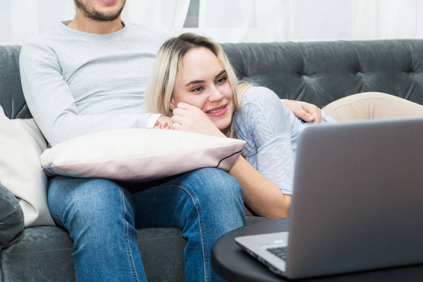 Young beautiful couple resting on the couch and watching online video from a laptop in the living room. - Fotografie, Obrázek