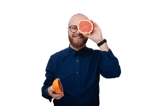 Photo of bearded holding two halves of grapefruit citrus fruit in hands, covering her eye with one over white background  - Valokuva, kuva
