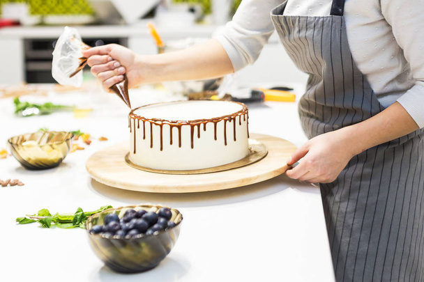 A confectioner squeezes liquid chocolate from a pastry bag onto a white cream biscuit cake on a wooden stand. The concept of homemade pastry, cooking cakes. - Φωτογραφία, εικόνα
