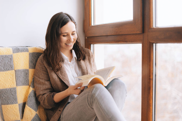 Photo of relaxed woman sitting on a couch near a window with a book. girl reading at home - Foto, Bild