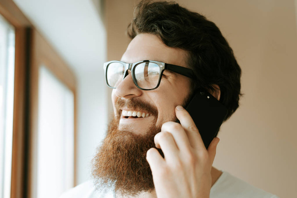 眼鏡をかけているひげを生やした男の写真は、家の窓の近くで携帯に立って話している  - 写真・画像