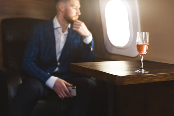 A young successful businessman in an expensive suit sits in the chair of a private jet with a glass of champagne in his hand and looks out the window - Foto, afbeelding