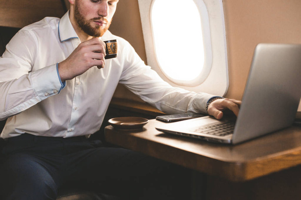 Attractive and successful businessman drinking coffee and working behind a laptop while sitting in a chair of his private jet. - Foto, Imagem